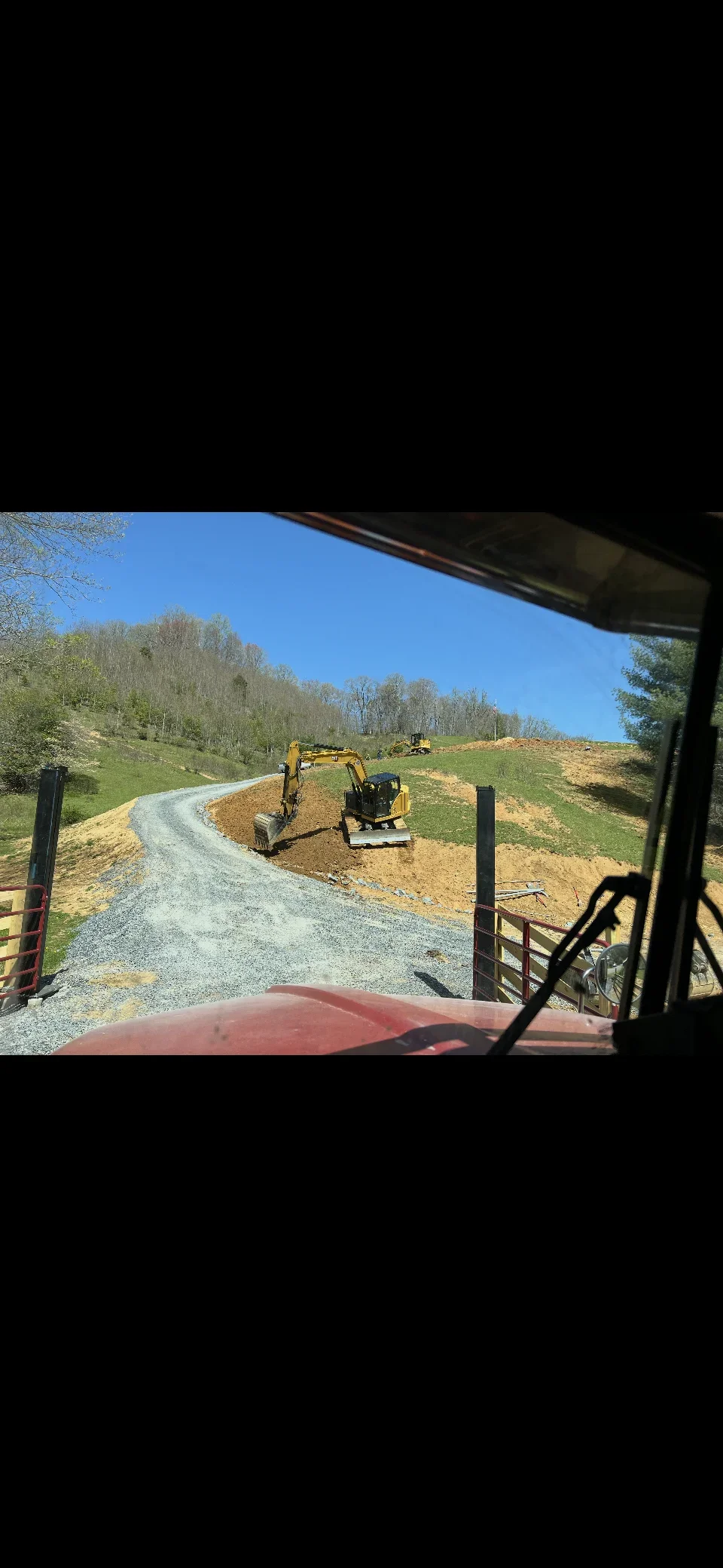 Compact excavator grading crushed stone driveway on sloped rural property with metal gates